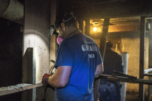 NECHAMA volunteer shines a headlamp onto a damaged wall in a basement, tools in hand to begin removing damaged wall.