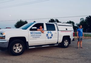 Nechama volunter smiles out the open window of a white pickup truck with the blue logo below him on the door. Another volunteer stands behind the truck smiling at something in the distance